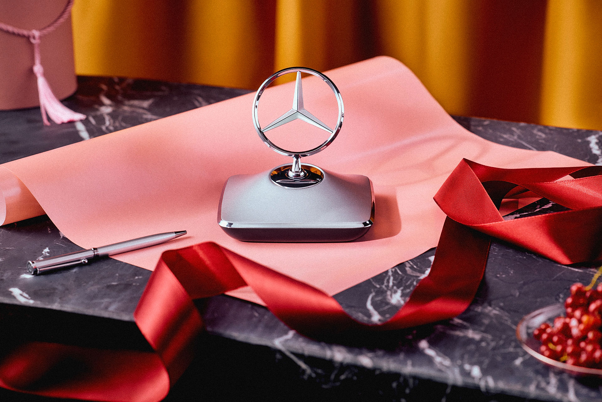 Mercedes-Benz Geschenkideen Gezeigt werden Mercedes-Benz Geschenkideen mit Briefbeschwerer in Sternform, Geschenkband und stilvoller Geschenkverpackung auf einem Marmortisch.