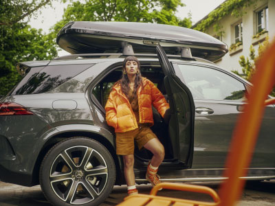 Mercedes-Benz Zubehör Trägersysteme Abgebildet ist eine junge Frau in orangener Jacke. Sie steht in der geöffneten Hintertür eine grauen Mercedes-Benz, welcher eine Mercedes-Benz Dachbox montiert hat.