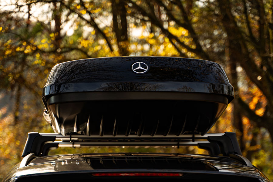 Abgebildet wird eine schwarze Mercedes-Benz Dachbox montiert auf dem zugehörigen Relingträger. Im Hintergrund sind unscharf Bäume in Herbstfarben zu sehen.