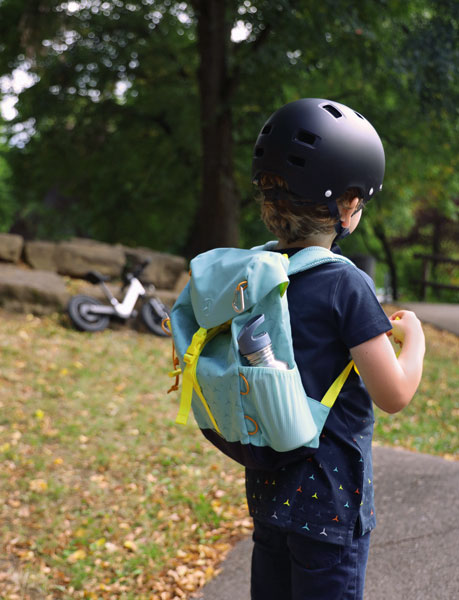 Zu sehen ist ein Kind von hinten, welches eine dunkel blaues Mercedes T-Shirt, einen schwarzen Helm und den türkisen Mercedes Kinderrucksack trägt.