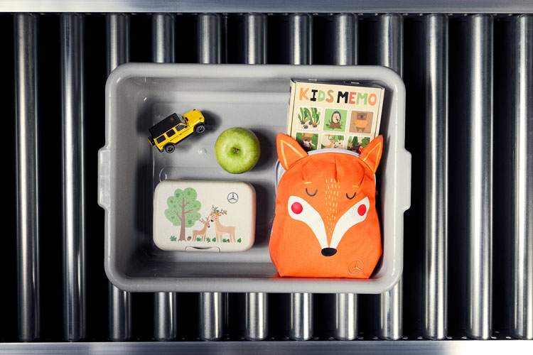 Abgebildet ist eine graue Plastikbox auf einem Rollband, in welcher neue Artikel der Mercedes-Benz Kinder Kollektion 