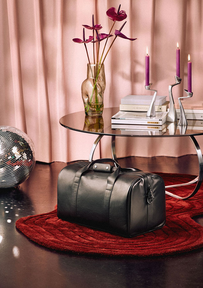 Mercedes-Benz Geschenkideen für Ihn Zu sehen ist eine schwarze Mercedes-Benz Reisetasche auf rotem Teppich vor modernem Wohnambiente als Geschenkidee für Ihn.