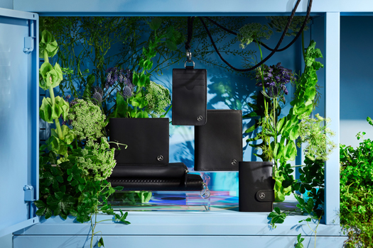Zu sehen sind verschiedene schwarze Lederwaren der Mercedes-Benz Collection. Die Brieftaschen sind vor einem bunten Hintergrund präsentiert - in einem hellblauen Regal mit grünen Pflanzen dekoriert.