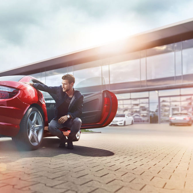 Zeigt einen Mann, welcher bei schönem Wetter neben seinem Auto kniet.