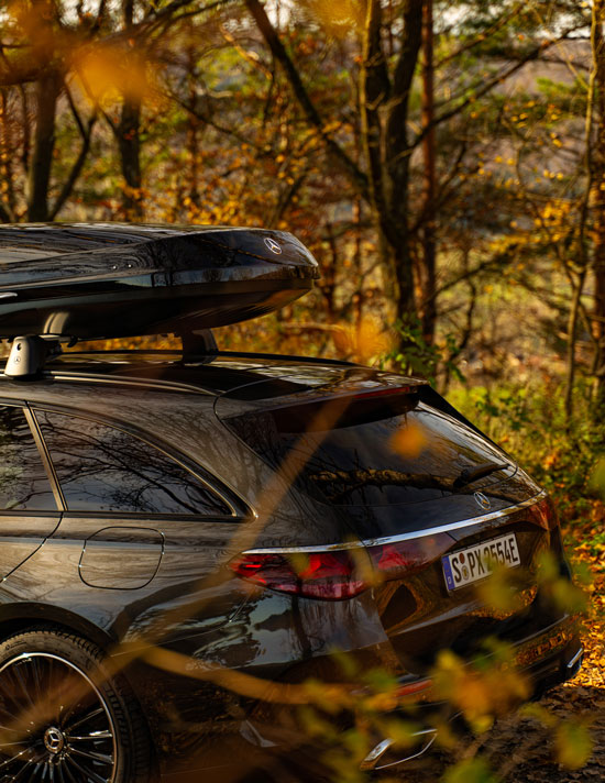 Mercedes-Benz Zubehör Abgebildet wird ein schwarzer Mercedes-Benz mit montierter Dachbox vor einem herbstlichen Waldhintergrund.